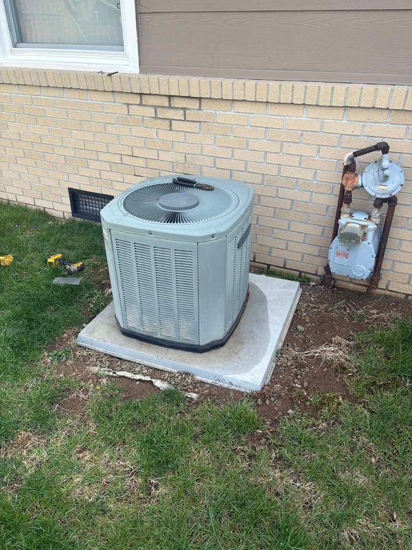 An outdoor AC unit on a concrete pad next to a brick house wall with a gas meter assembly nearby.
