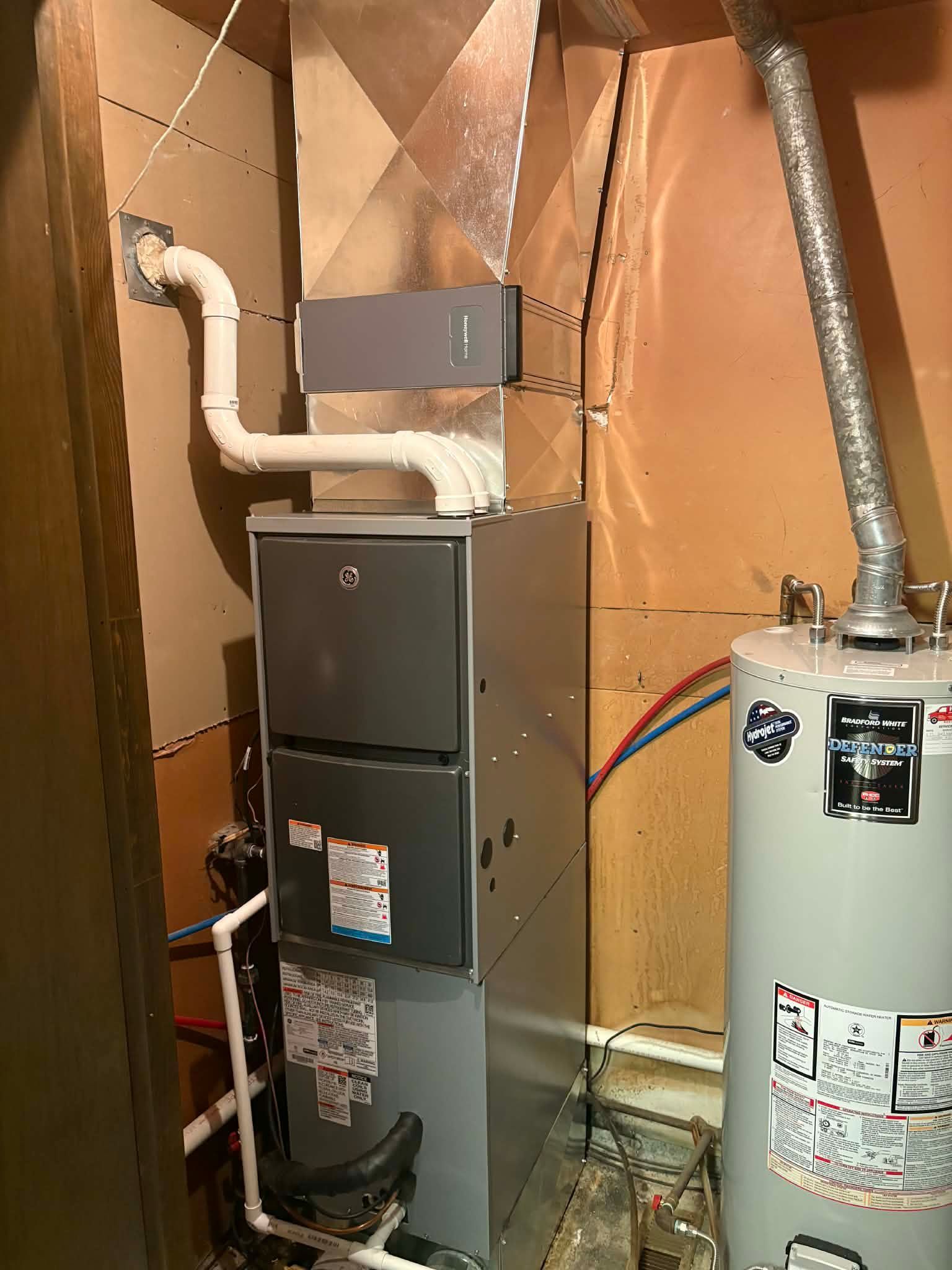 A gray furnace unit with white PVC venting and a metal duct, positioned next to a water heater in a utility closet.
