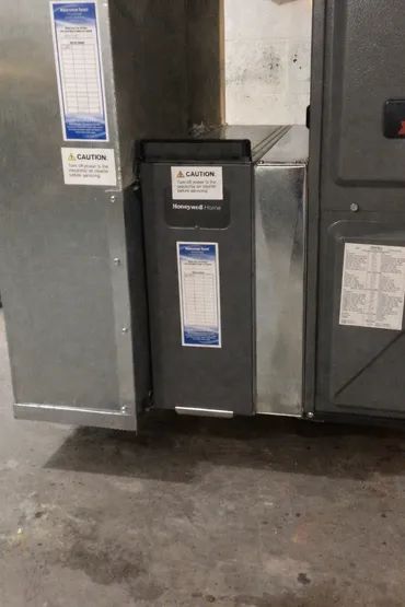 A gray furnace air cleaner unit installed between two metal ductwork sections in a utility room.