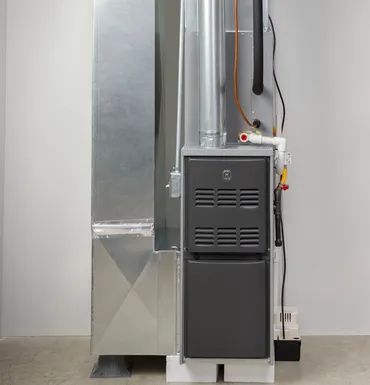 A vertical gas furnace unit with metallic ductwork, white condensation piping, and a condensate pump on a gray floor.