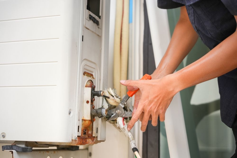 A technician uses pliers to work on the external copper pipe connections of a white air conditioning unit.