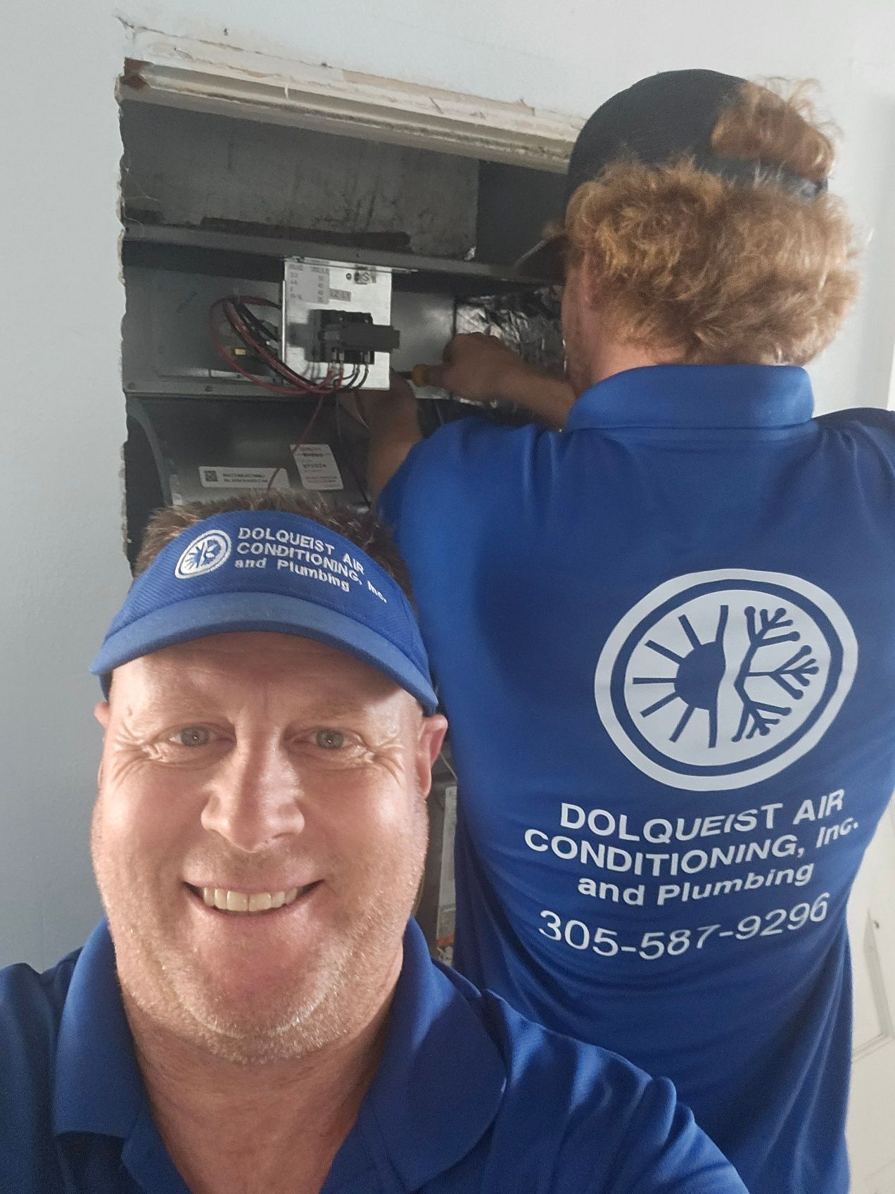 Two people in blue shirts, one taking a selfie, the other working on an air conditioning unit.