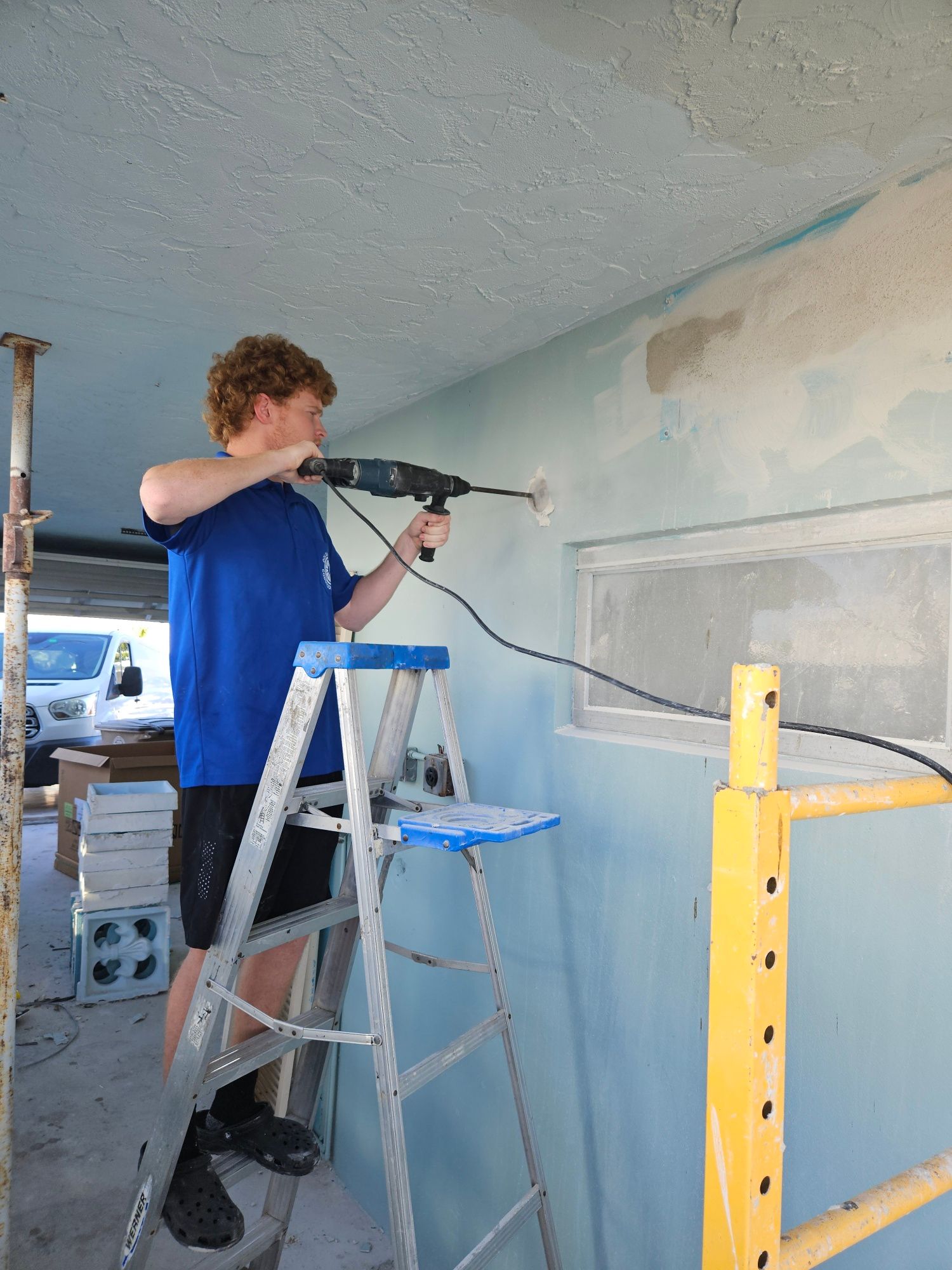 Man drilling into a light blue wall while standing on a stepladder. A window and scaffolding are present.