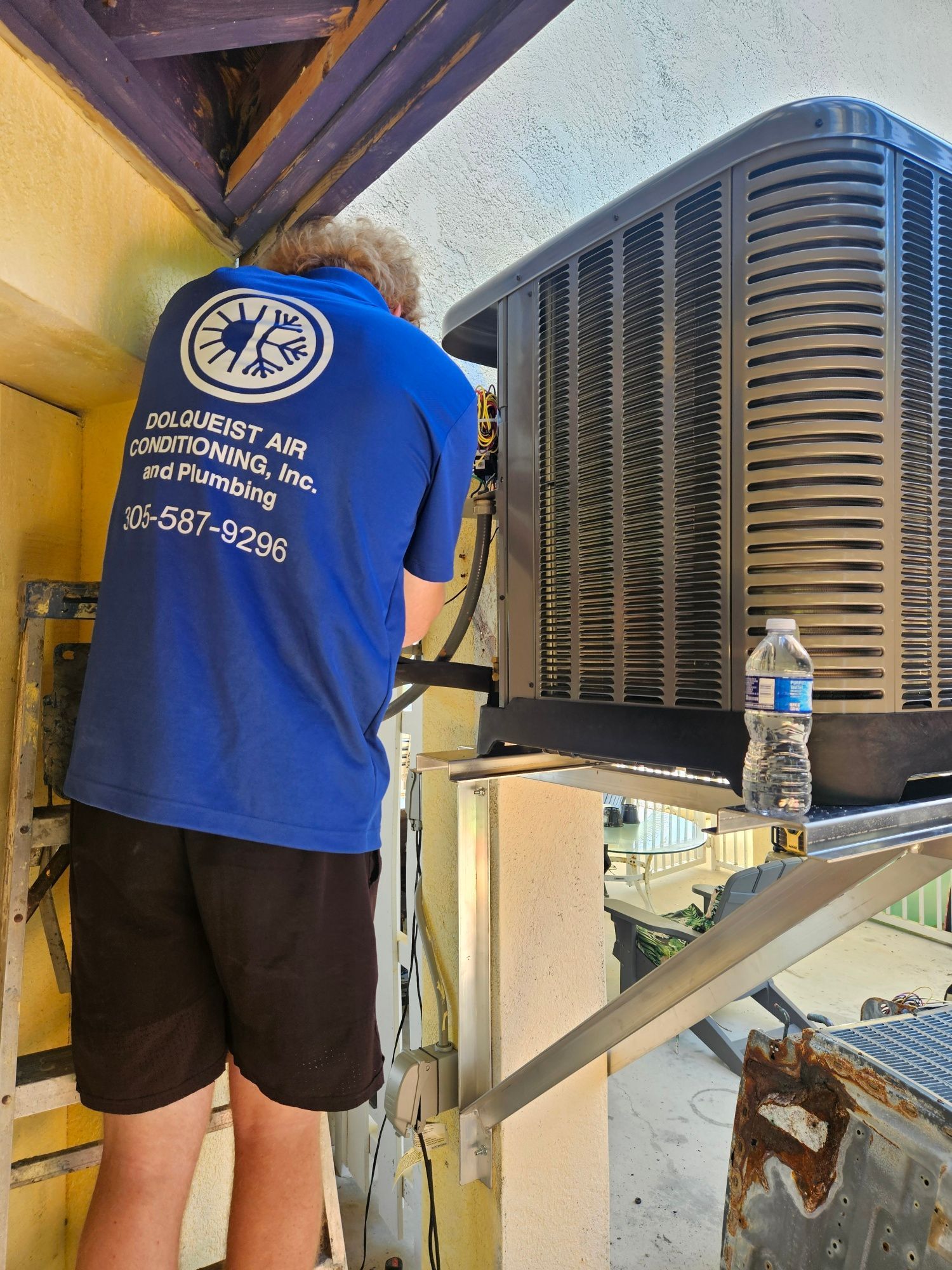 Person in blue shirt working on an AC unit outdoors. A water bottle is visible.