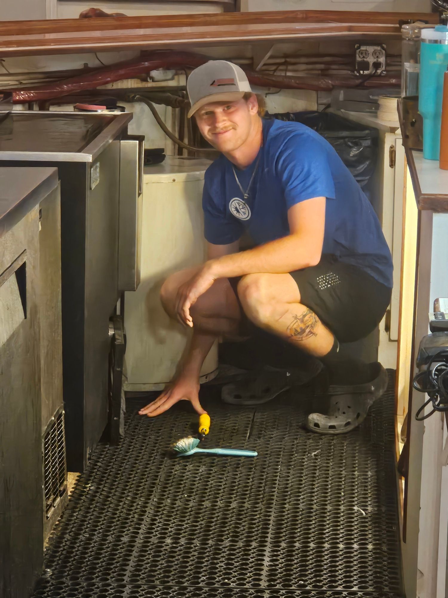Man crouches near a floor grate in a small kitchen, smiling and pointing toward the floor.