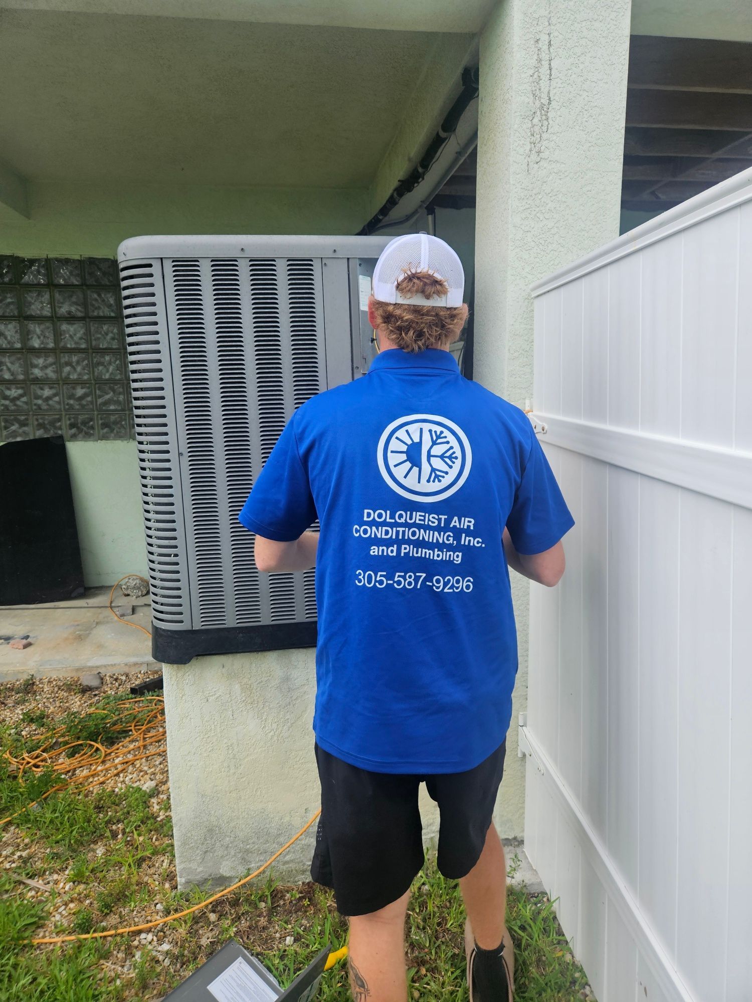 Person in blue shirt working on AC unit next to white fence.