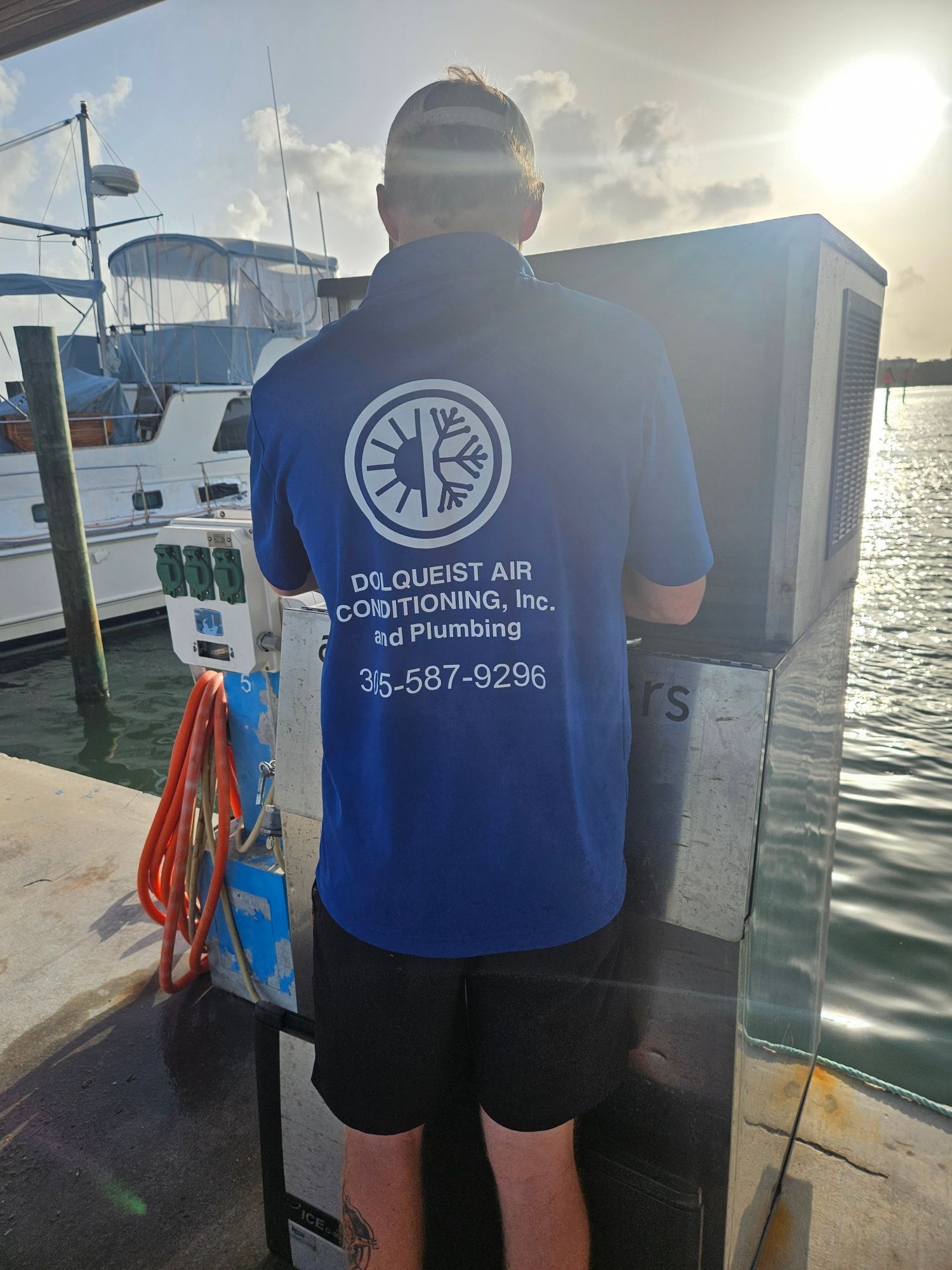 Man in blue shirt, working on machinery near water. Company logo visible. Bright sun, boats in background.