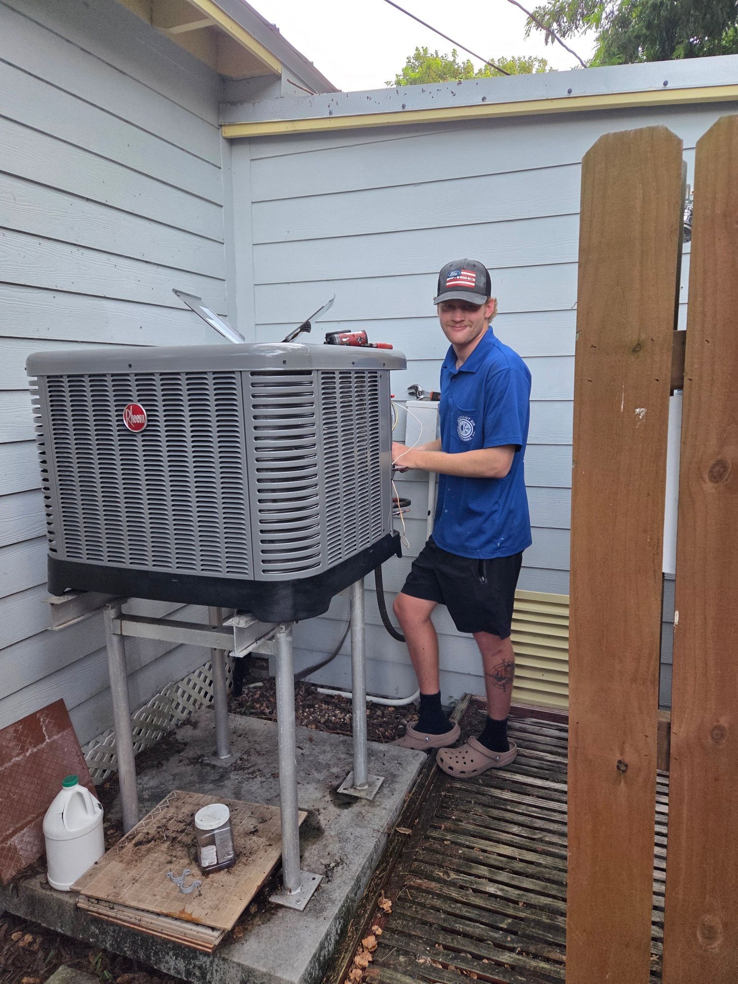 Man in blue shirt, shorts near a large outdoor air conditioning unit, outdoor setting.