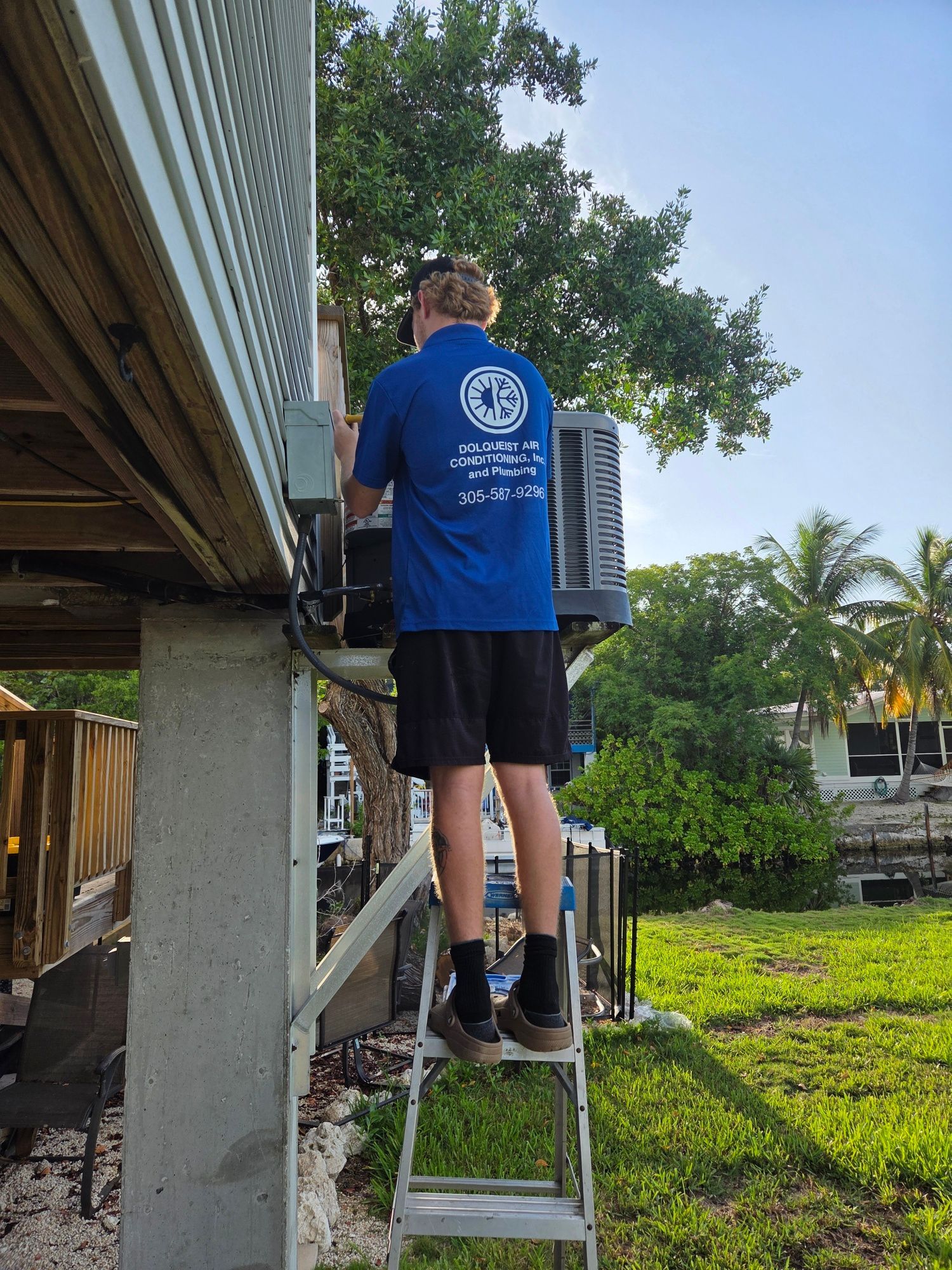 Man on ladder installing outdoor air conditioning unit under wooden deck.