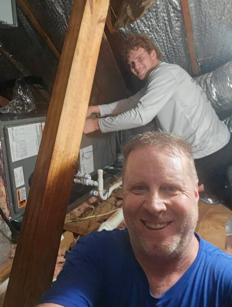 Two men in an attic working on HVAC equipment. One takes a selfie, both smiling.