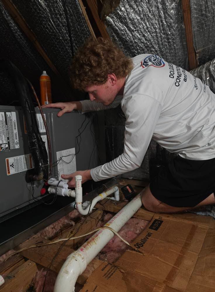 Person inspecting HVAC unit in an attic. Leaning forward, hand on the unit, surrounded by ductwork.