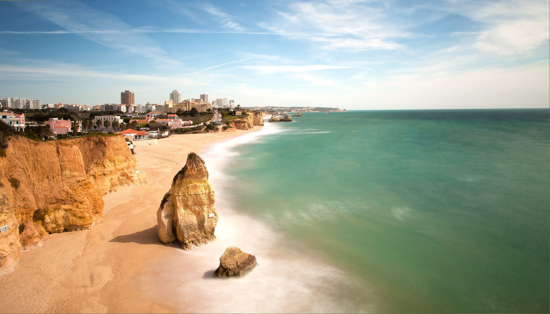 Cliffs and beach with turquoise water and a city in the background under a blue sky.