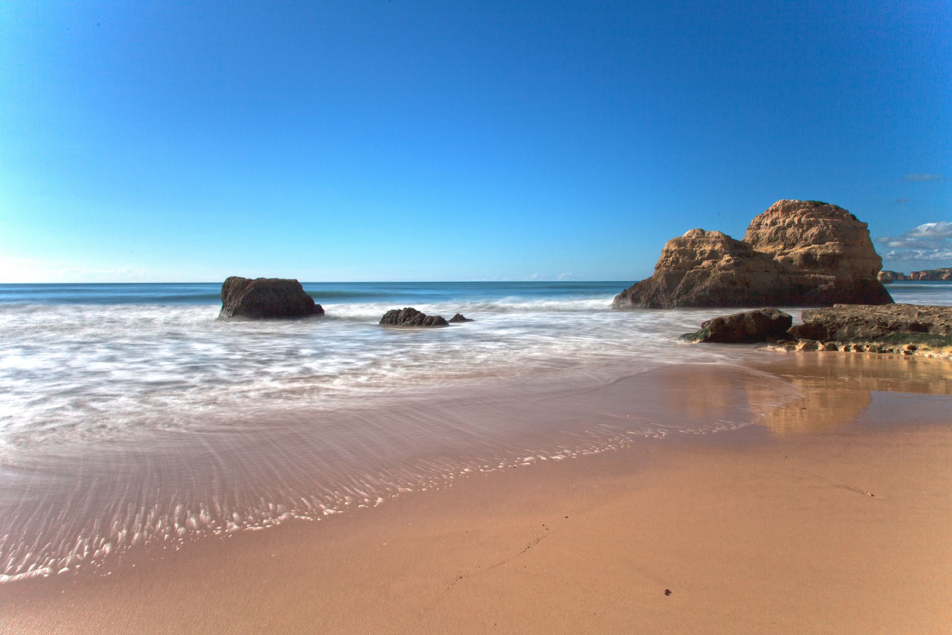 Sandy beach with clear blue sky and waves crashing around rocky outcrops.