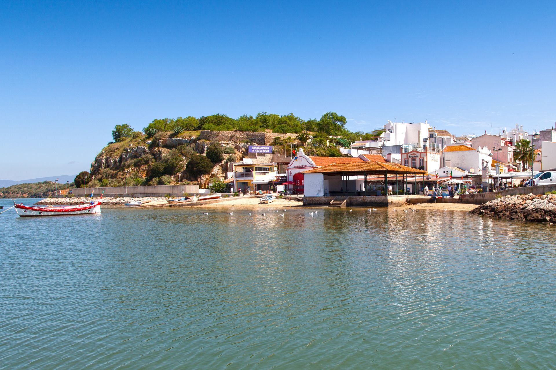 Coastal town scene; calm blue water, small boats, buildings with white and red accents, and a hill with green foliage.