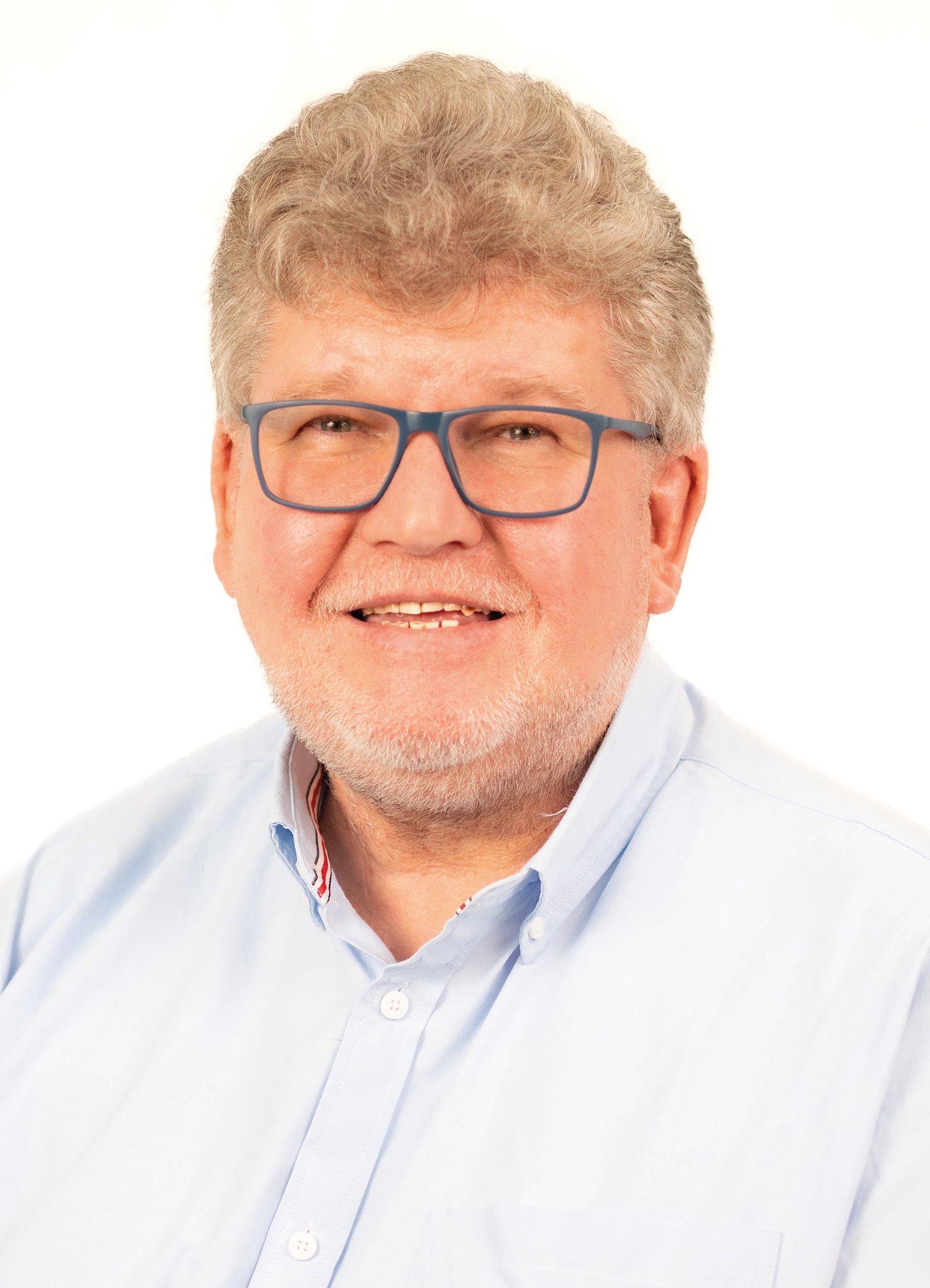 Mann mit Brille, lächelnd, trägt ein hellblaues Hemd. Graue Haare und grauer Bart. Weißer Hintergrund.