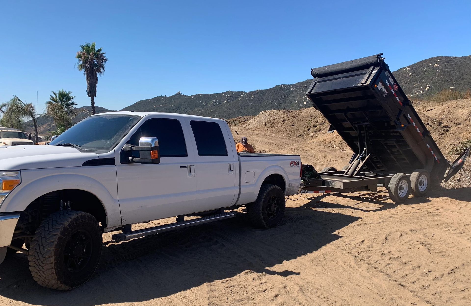White pickup truck towing a dump trailer on dirt road, mountains in the background, sunny day.