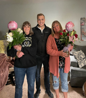 A man and two women are holding flowers and balloons in a living room.