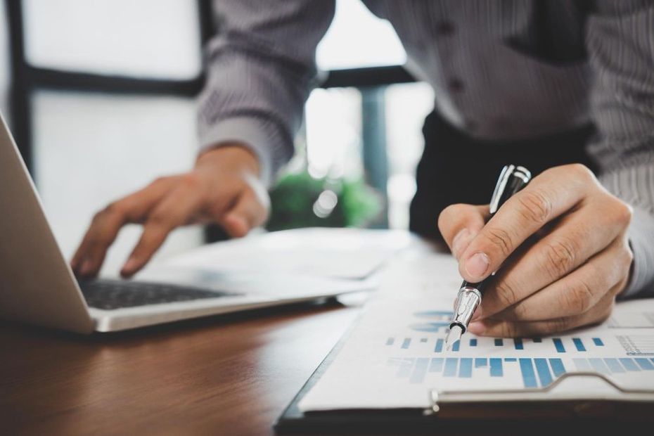 A person in a professional shirt points a pen at a paper bar chart while working on a laptop at a desk.