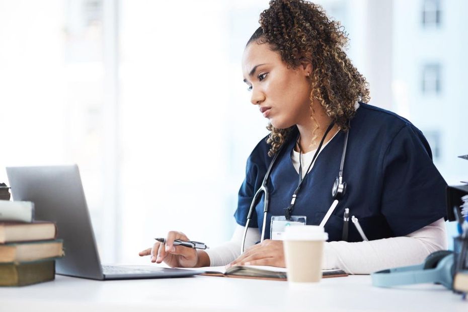 A medical professional wearing blue scrubs and a stethoscope uses a laptop while sitting at a desk with books and coffee.