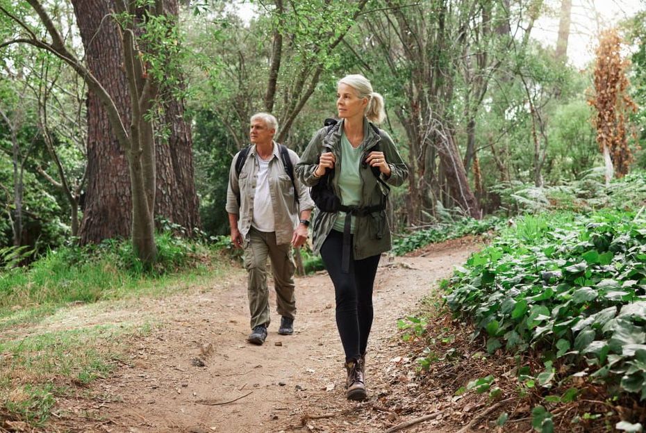 Two hikers with backpacks walk along a dirt path through a lush, green forest.