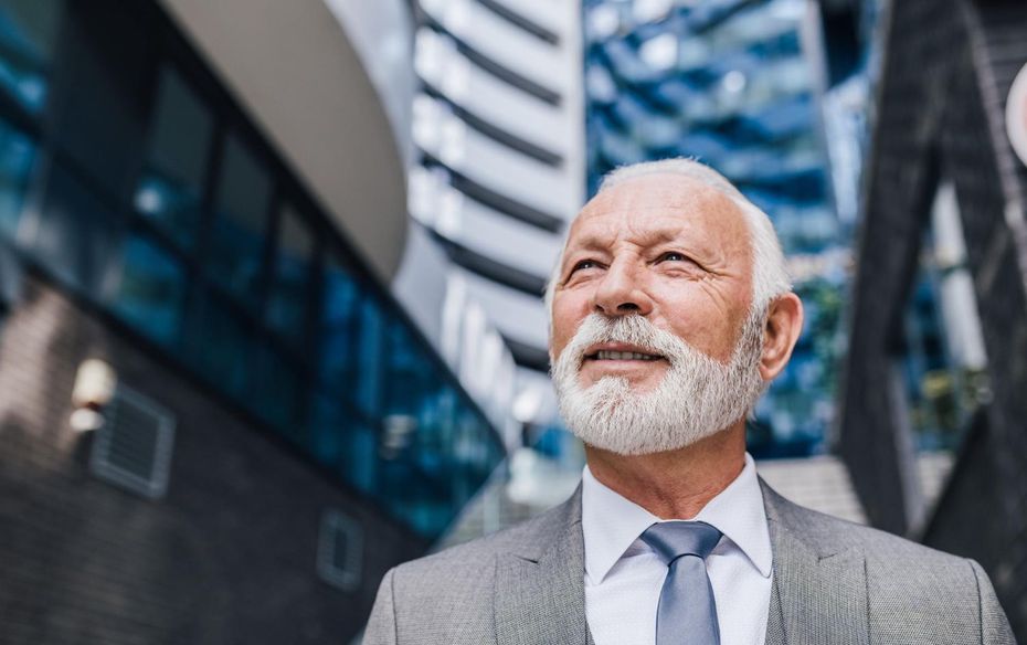 A person with a white beard wearing a gray suit and tie, looking upward against a background of modern office buildings.