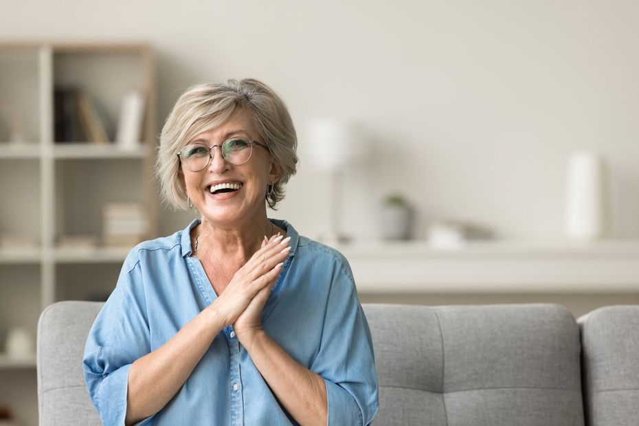 A smiling person in a blue shirt claps their hands together while sitting on a gray couch in a bright room.