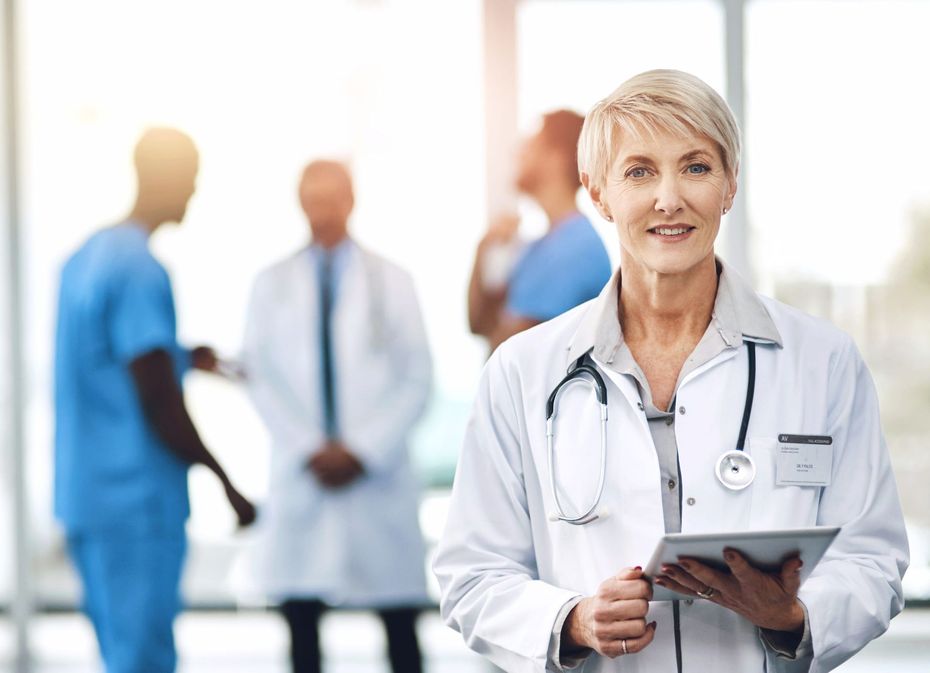 A medical professional in a white coat with a stethoscope holds a tablet in a hospital lobby with colleagues behind.