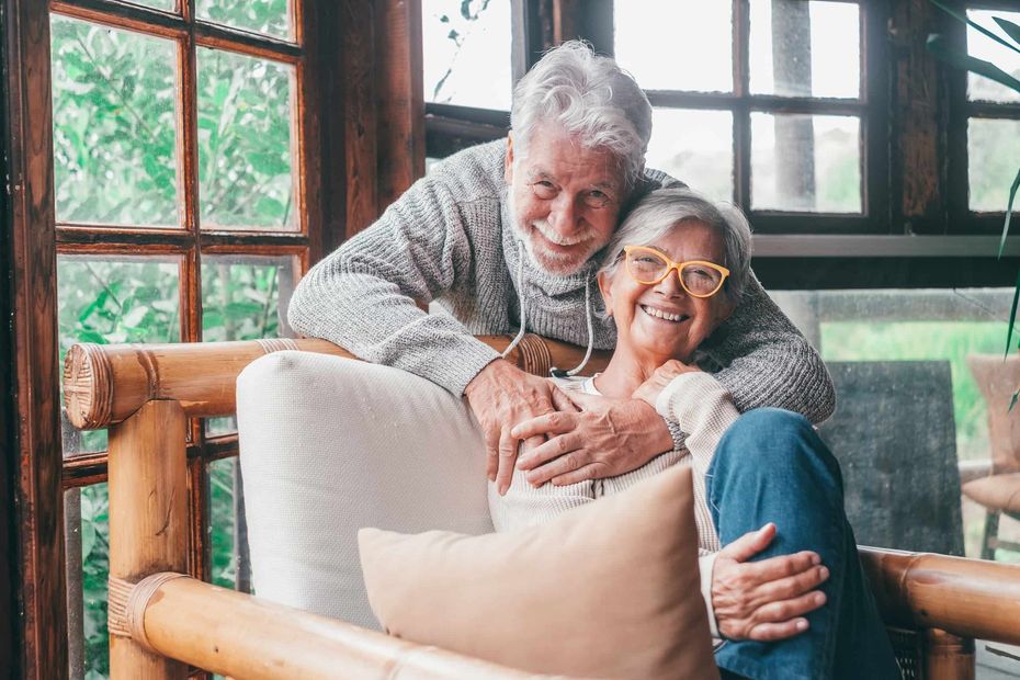 Two smiling people sitting together on a wooden chair in a sunlit room with large windows.