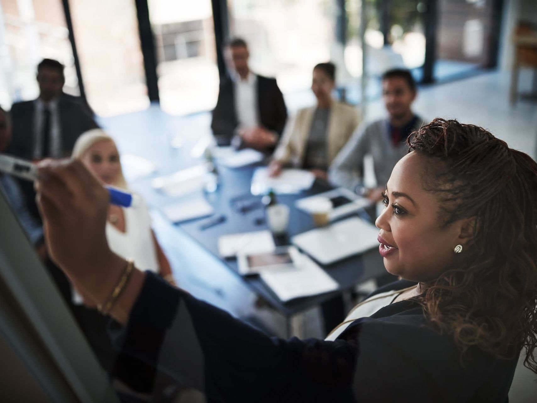 Woman writing on a whiteboard during a meeting; colleagues seated around a conference table.