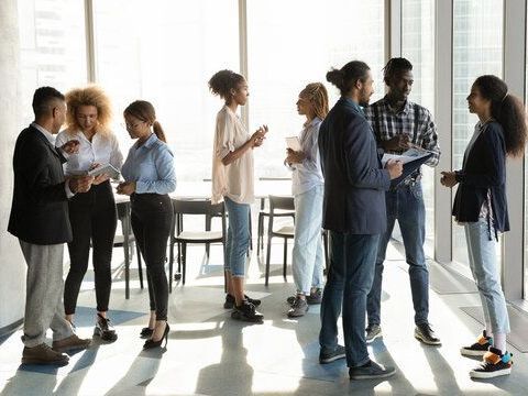 People in business attire talking and reviewing papers in a brightly lit office near a window.