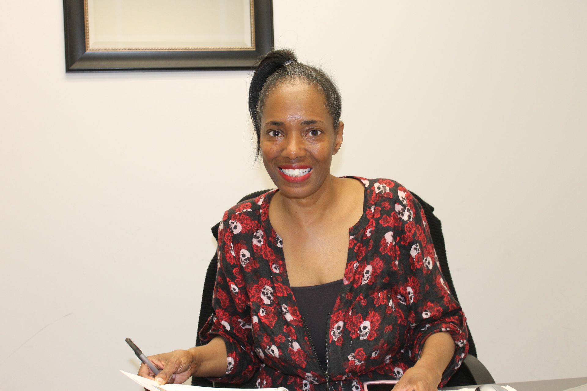 A woman is sitting at a desk with a pen in her hand and smiling.