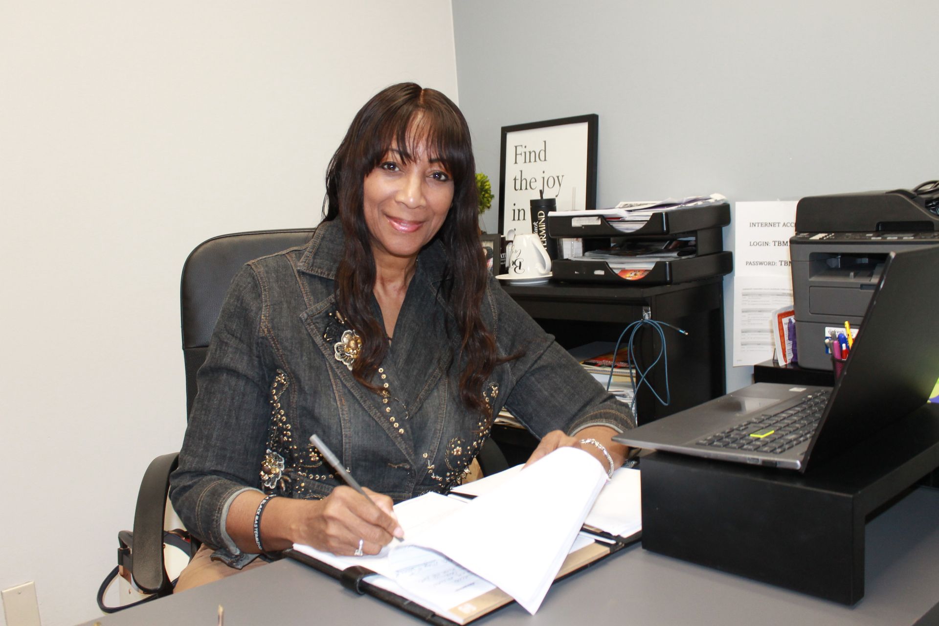 A woman is sitting at a desk with a laptop and a clipboard.