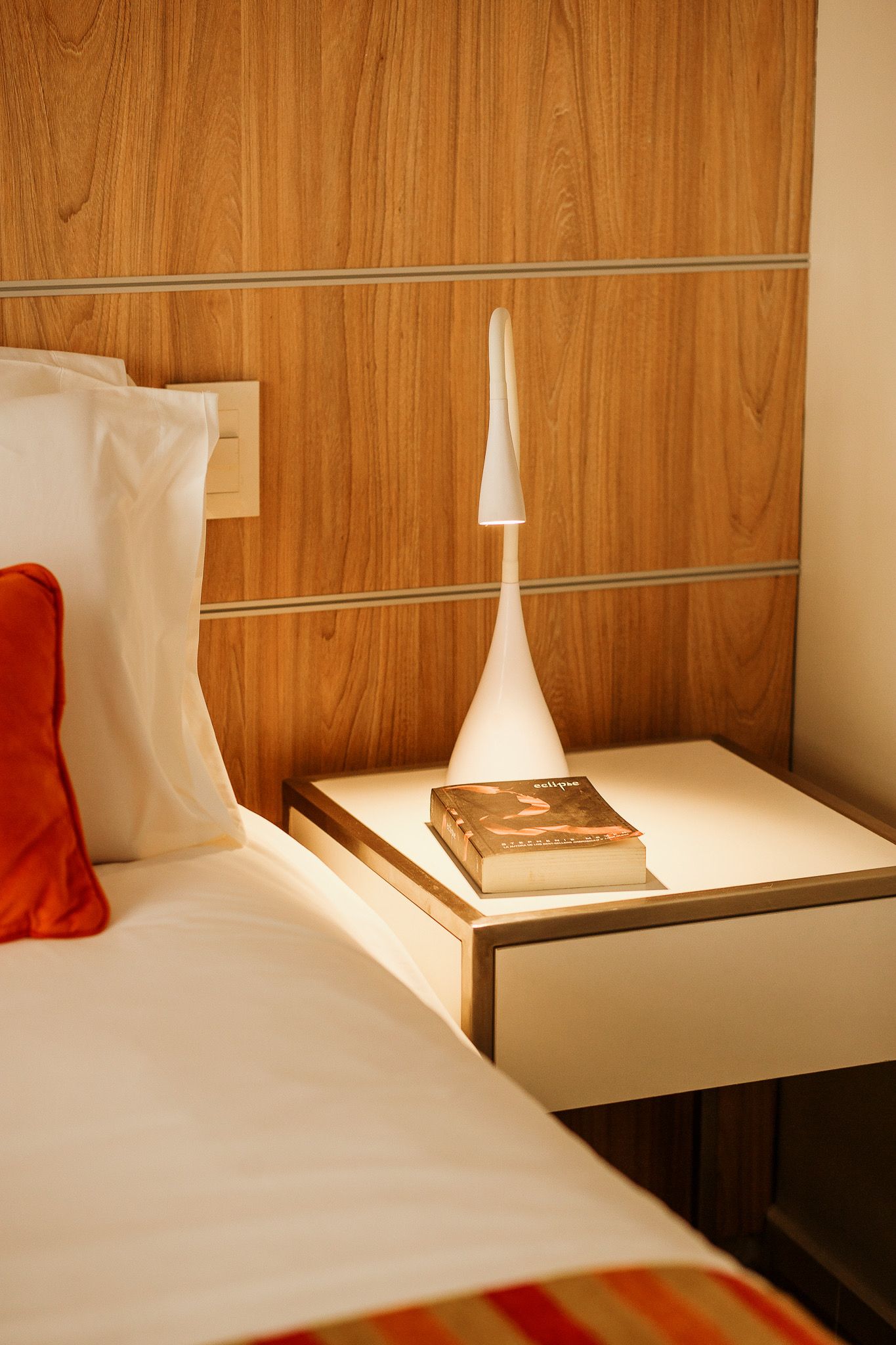 Bedside table with lamp and book next to a bed with white sheets and an orange pillow.