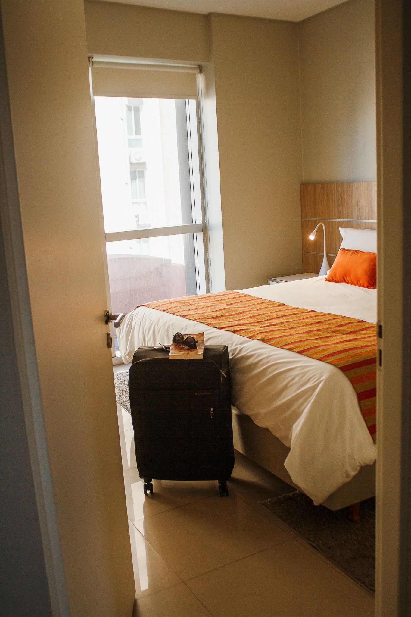 Bedroom interior with a bed, luggage, and window; sunlight illuminates the room.