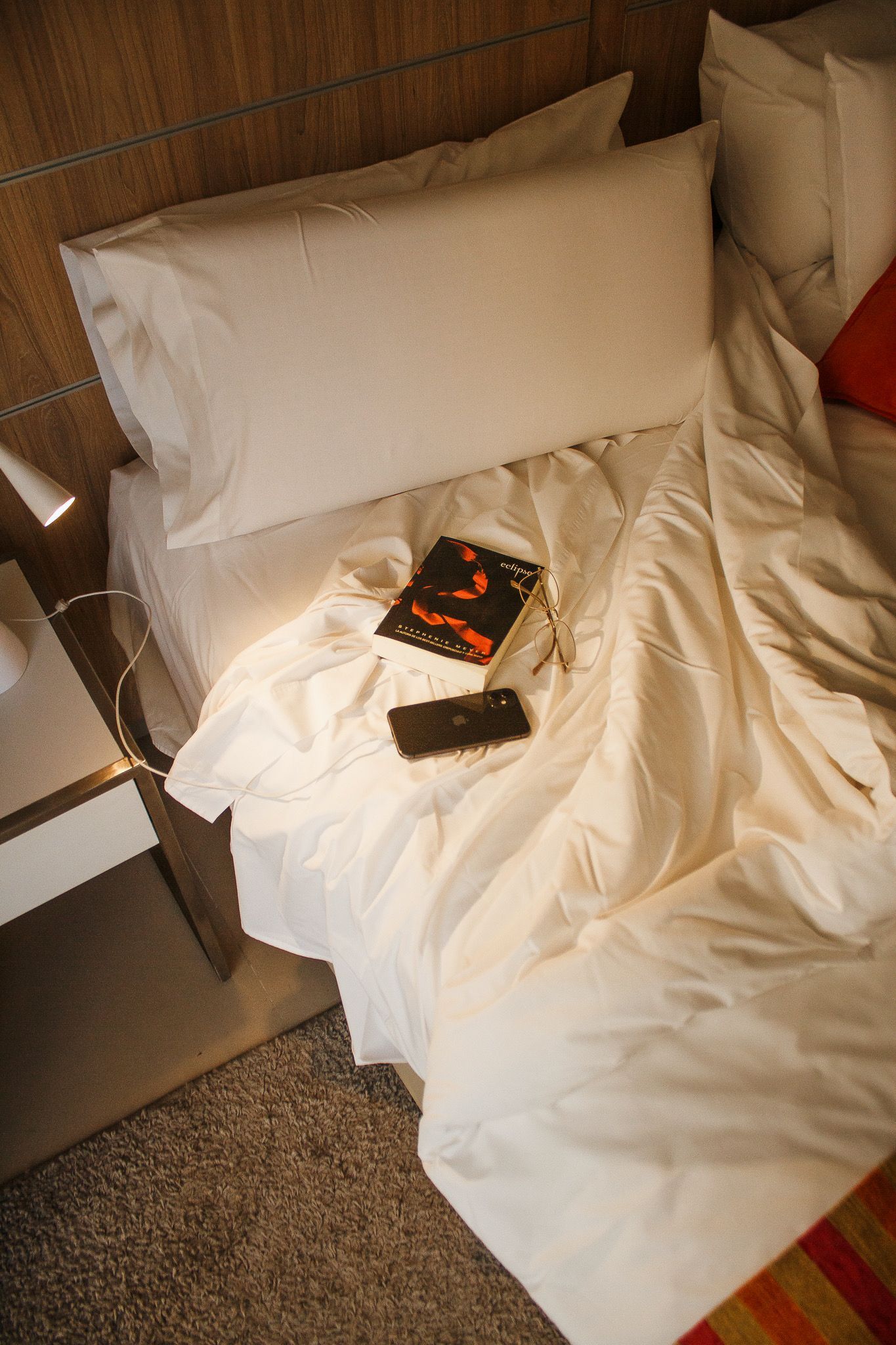 Bed with white sheets, pillow, book, and remote, partially unmade. Orange and red accents.
