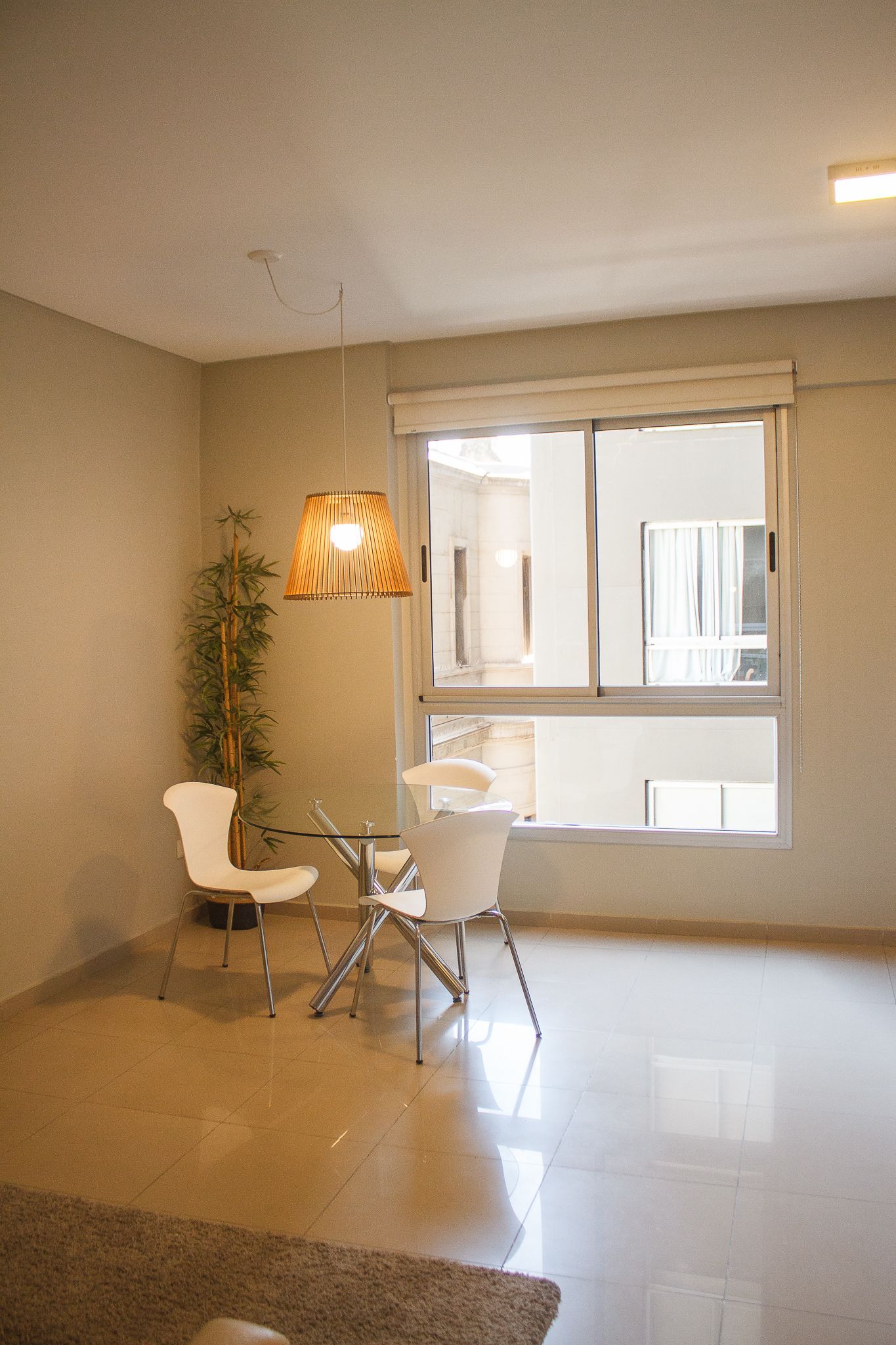 Empty interior: chairs, small table, window, tall plant, hanging light. Beige walls, shiny floor.