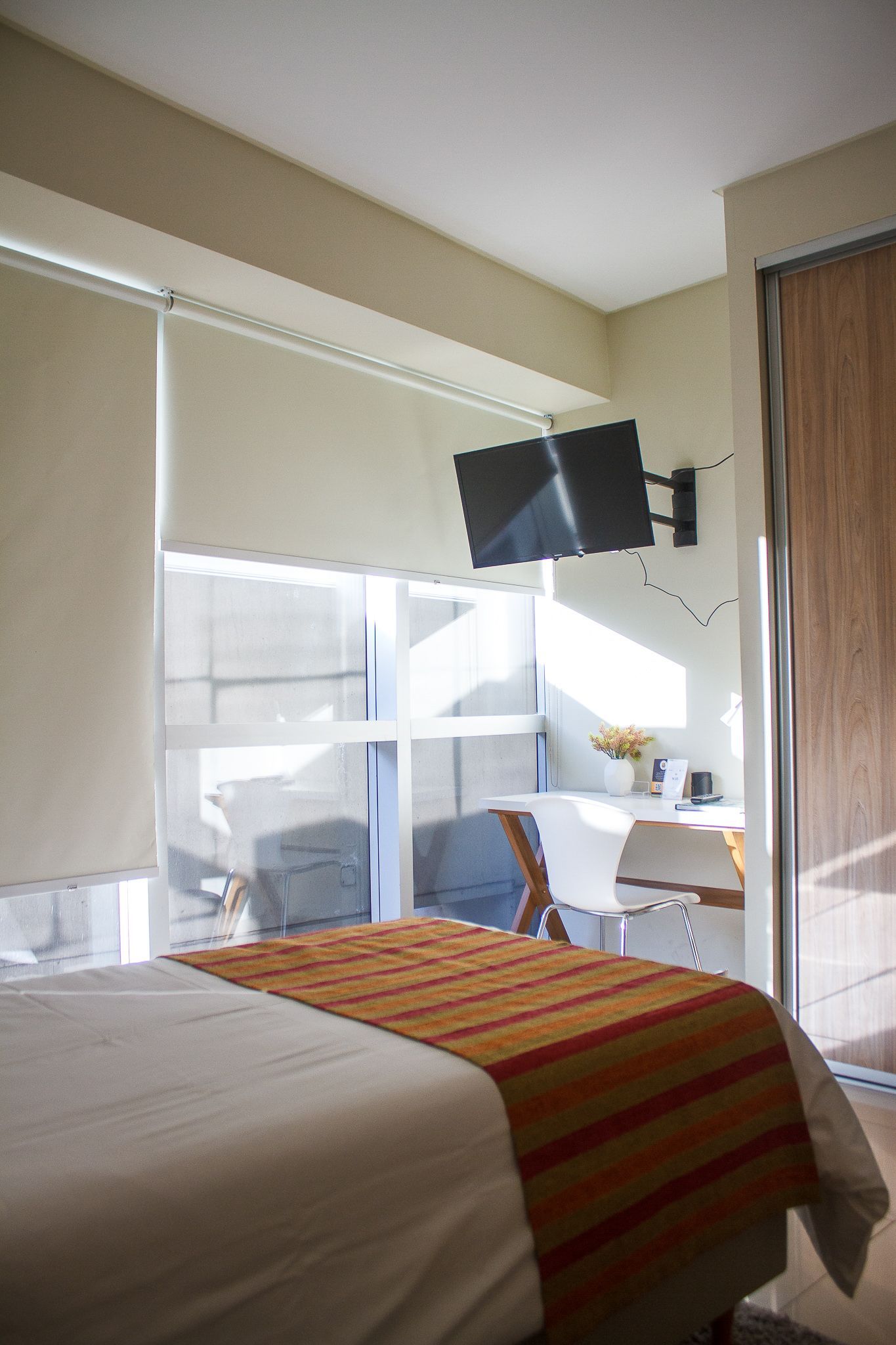Bedroom with bed, desk, window, and wall-mounted TV. Orange and brown bed runner.