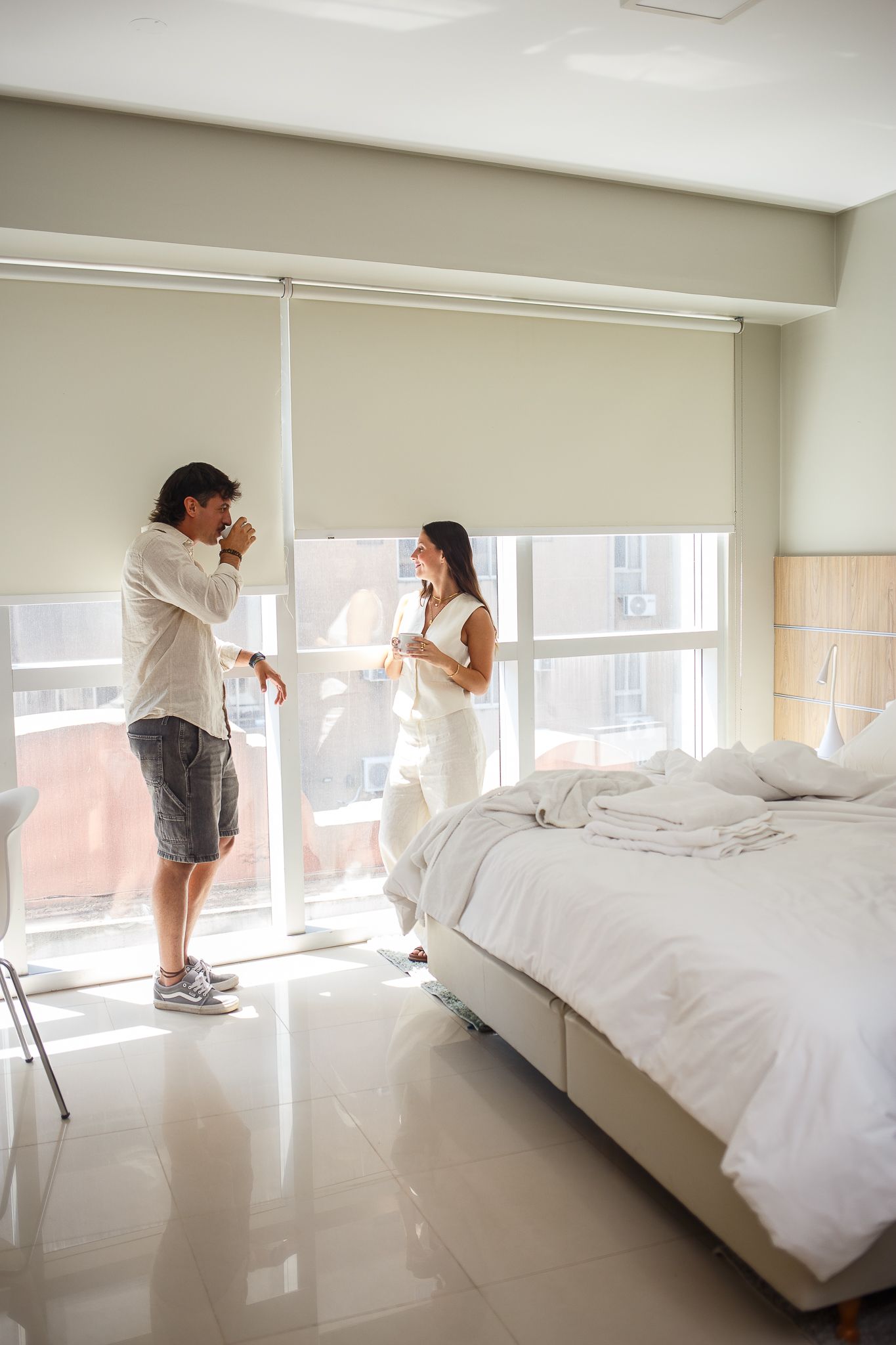 Couple near window, bedroom. Man gestures, woman holds glass. Bed unmade, bright light.