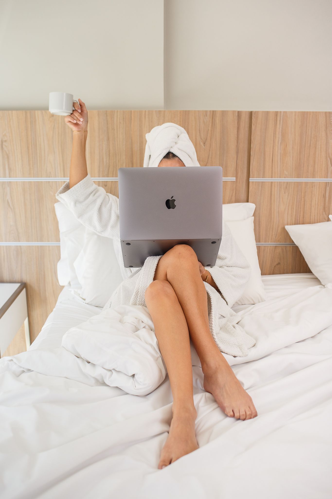 Woman in bathrobe with laptop, holding coffee cup in bed.