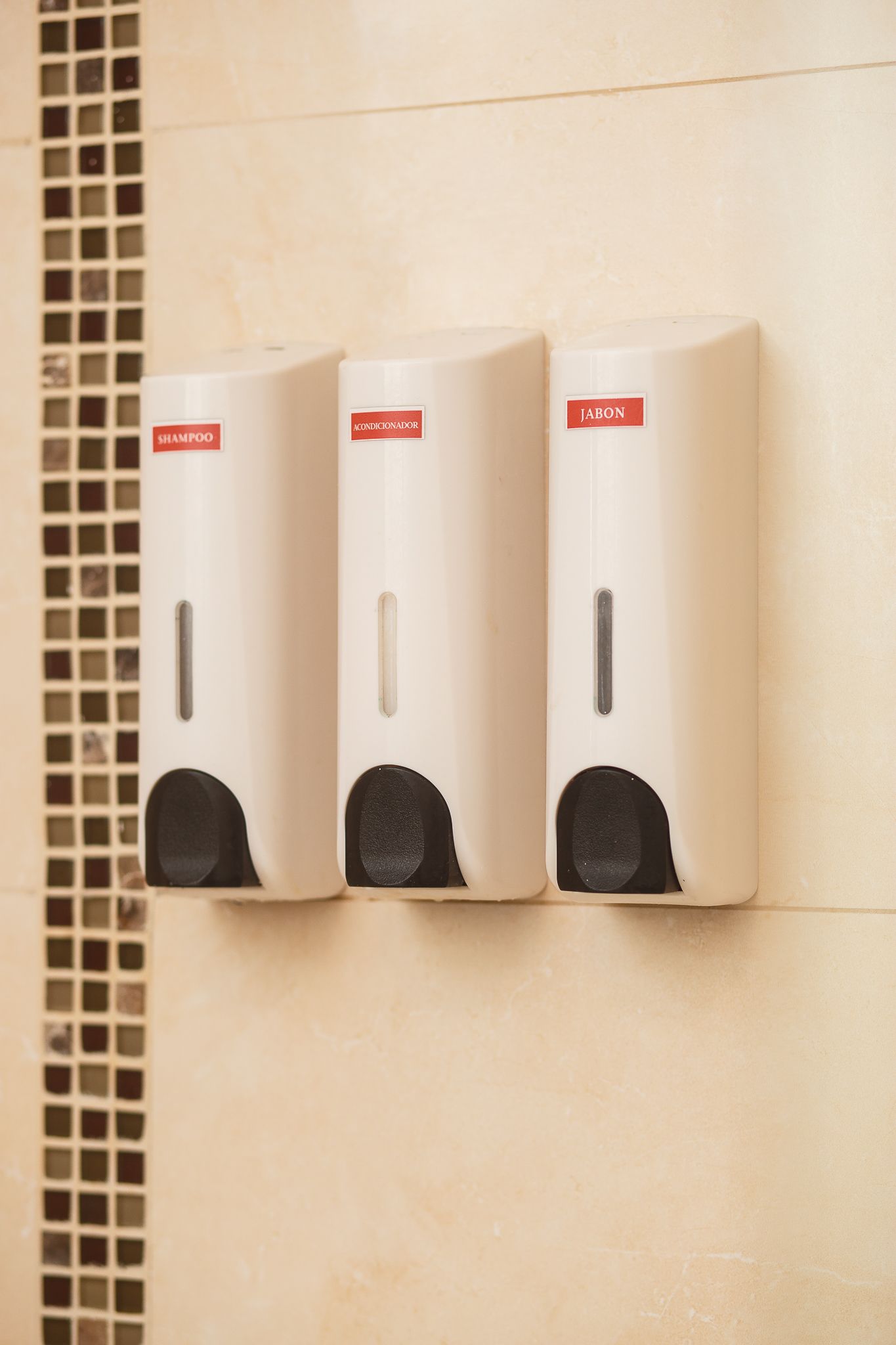 Three white soap dispensers mounted on a tiled wall, with a mosaic tile border on the left.