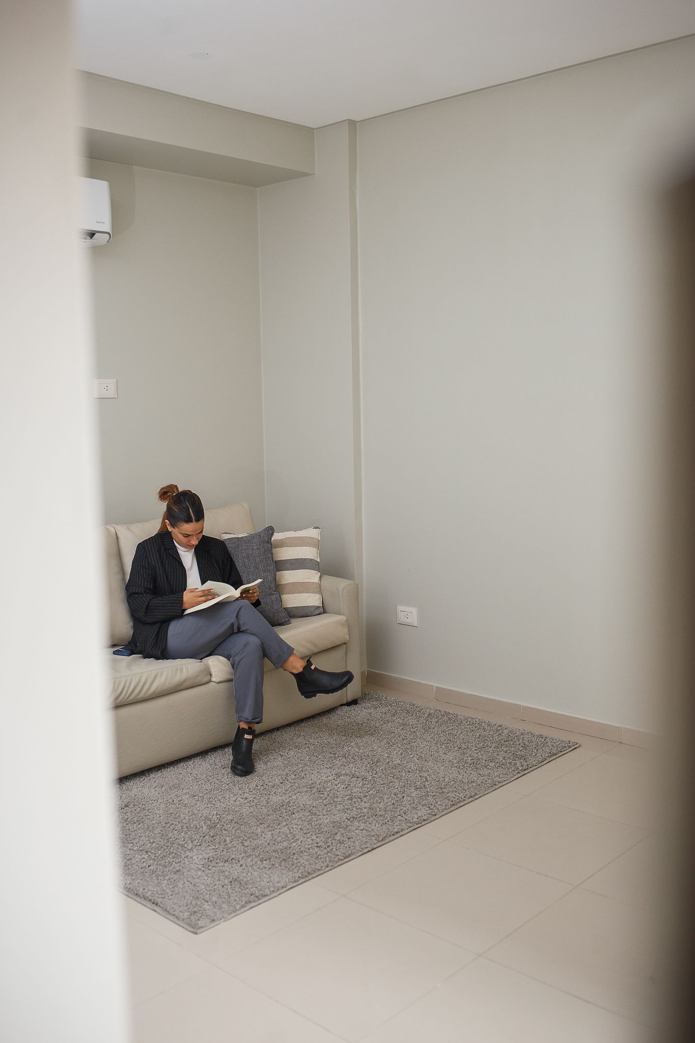Woman sitting on a beige couch, reading a book in a room with light grey walls and a rug.