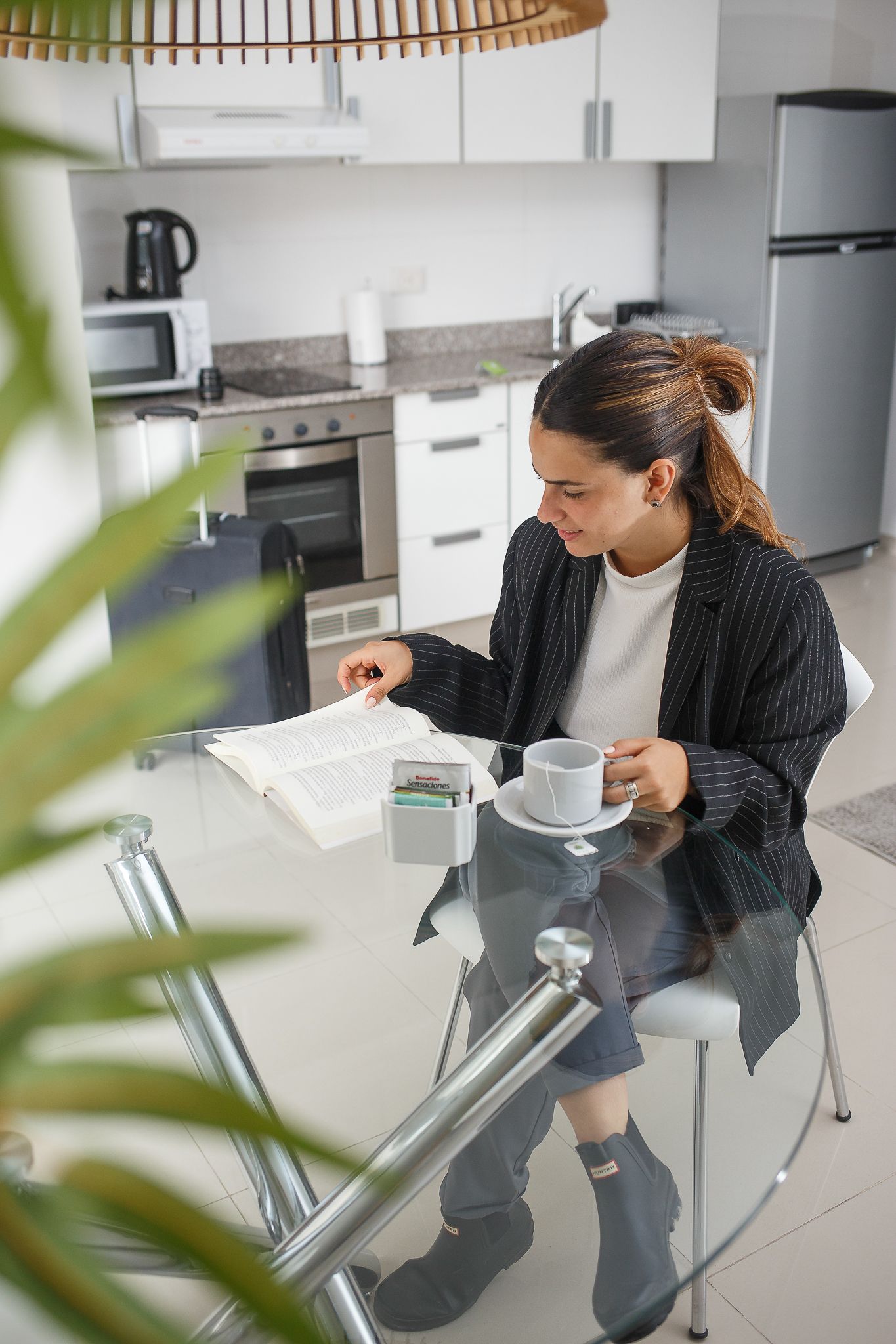 Woman sits at glass table, reading papers, holding a coffee cup in a kitchen.