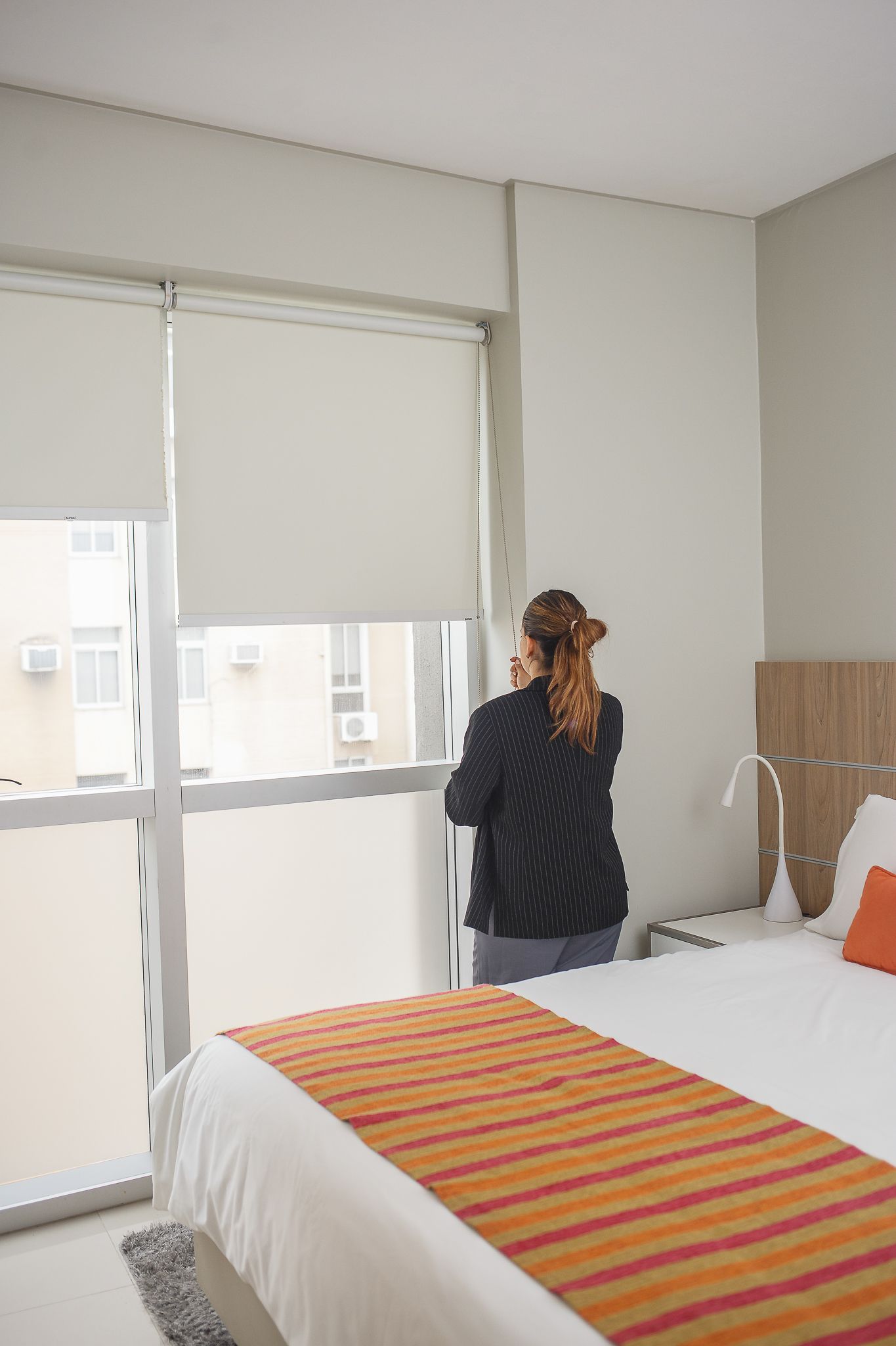 Woman looking out window in a bedroom with closed blinds; bed with orange and red runner.