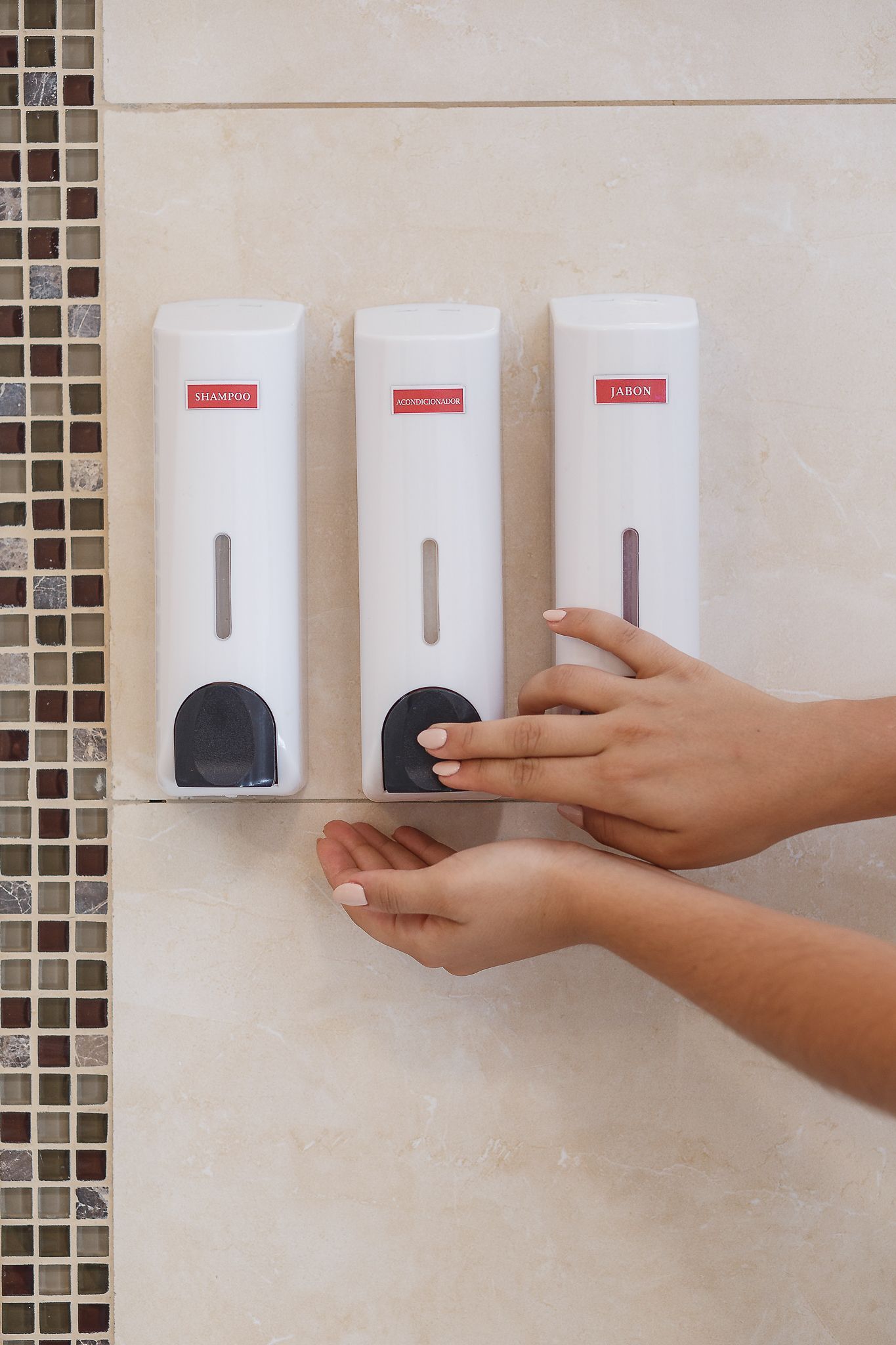 Three white soap dispensers mounted on a tiled wall, a hand presses one.