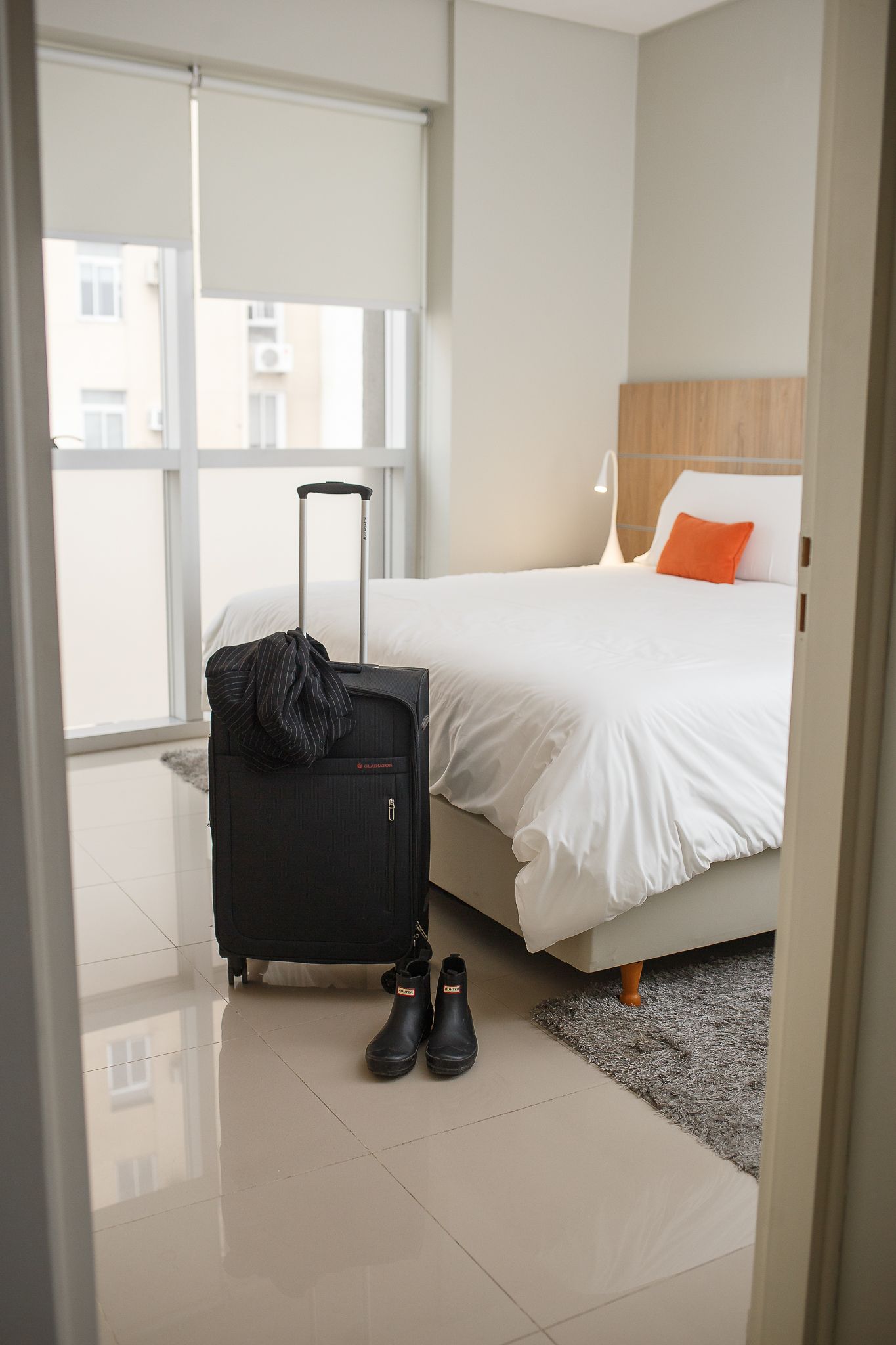 A bedroom with a bed, luggage, and shoes near the door. Window with blinds, light gray walls.