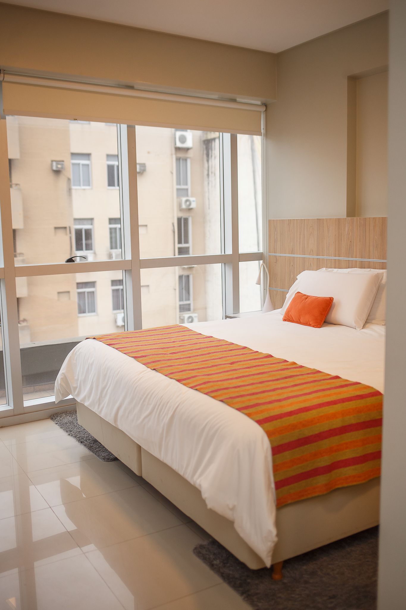 Bedroom with floor-to-ceiling windows overlooking a city, bed with orange accents, and light flooring.