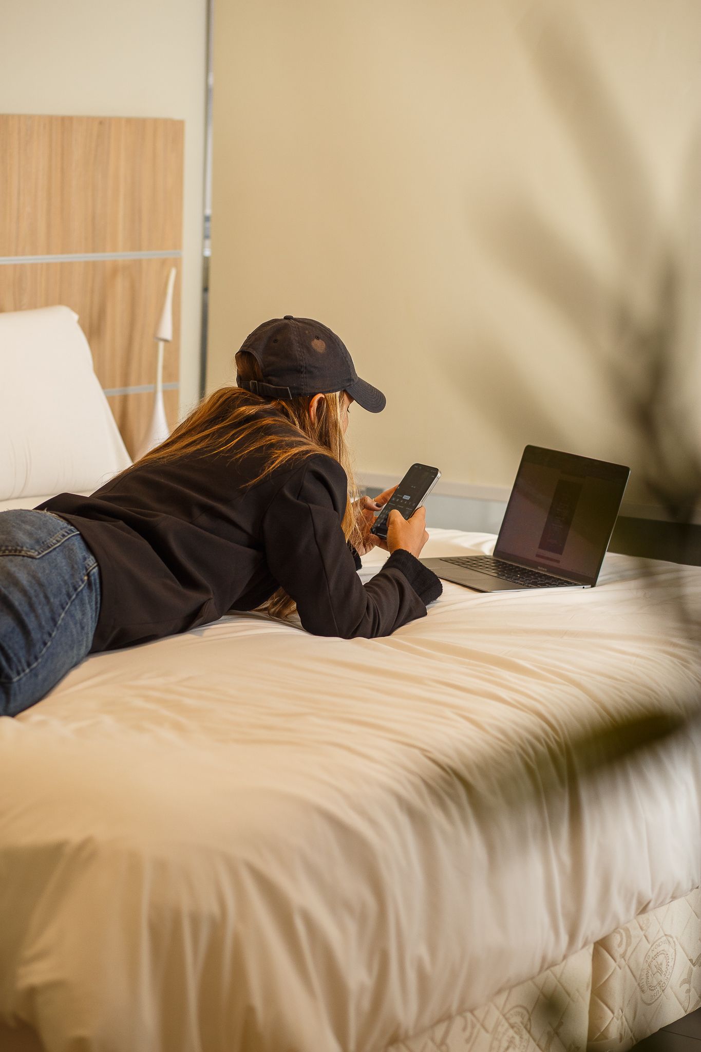 Person lying on bed using a laptop and phone, wearing a black cap. Neutral setting with cream-colored bedding.