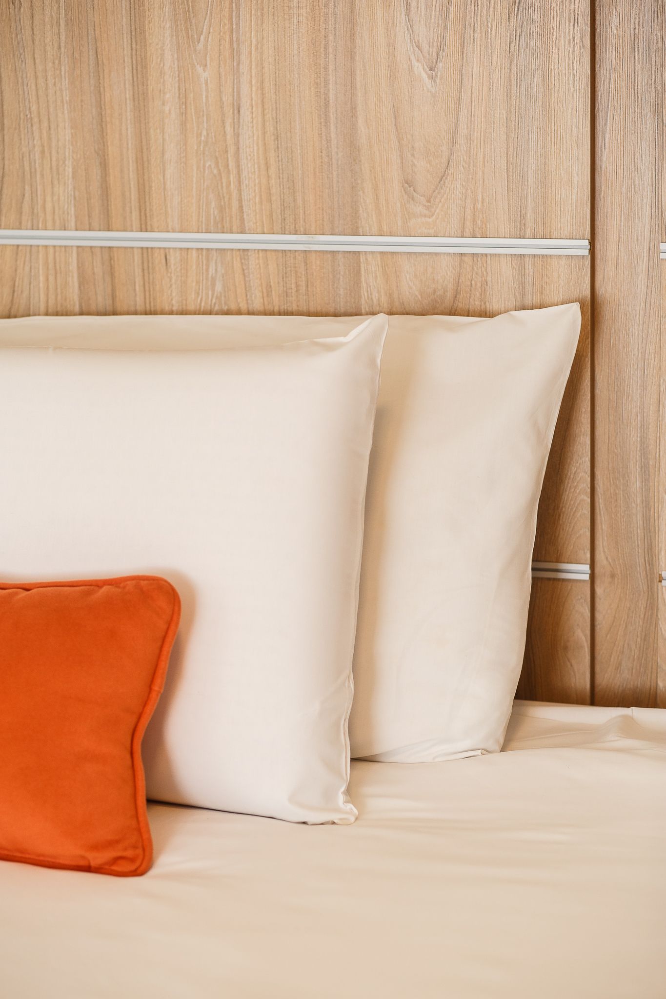 Bed with white pillows and orange decorative pillow against a wood headboard.