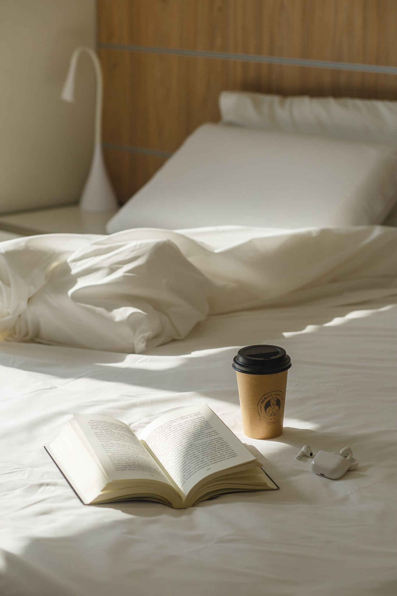 Open book, coffee cup, and earbuds on a bed with rumpled white sheets; a lamp is visible in the background.