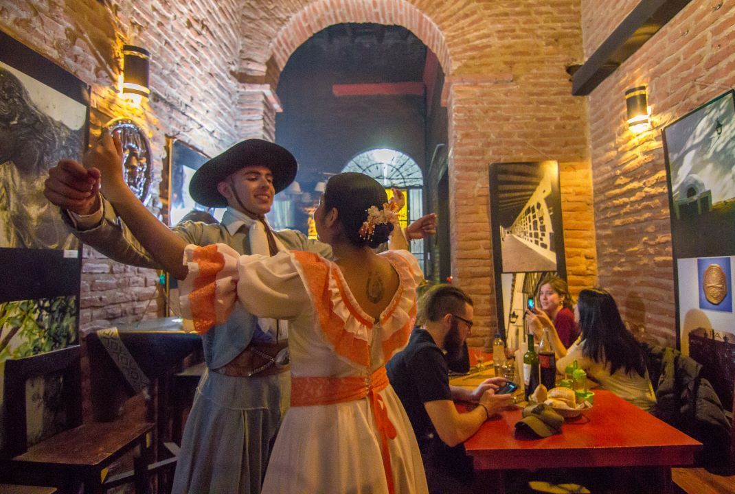 Couple in traditional dress dancing in a brick-walled bar, lit with warm light; patrons watch.